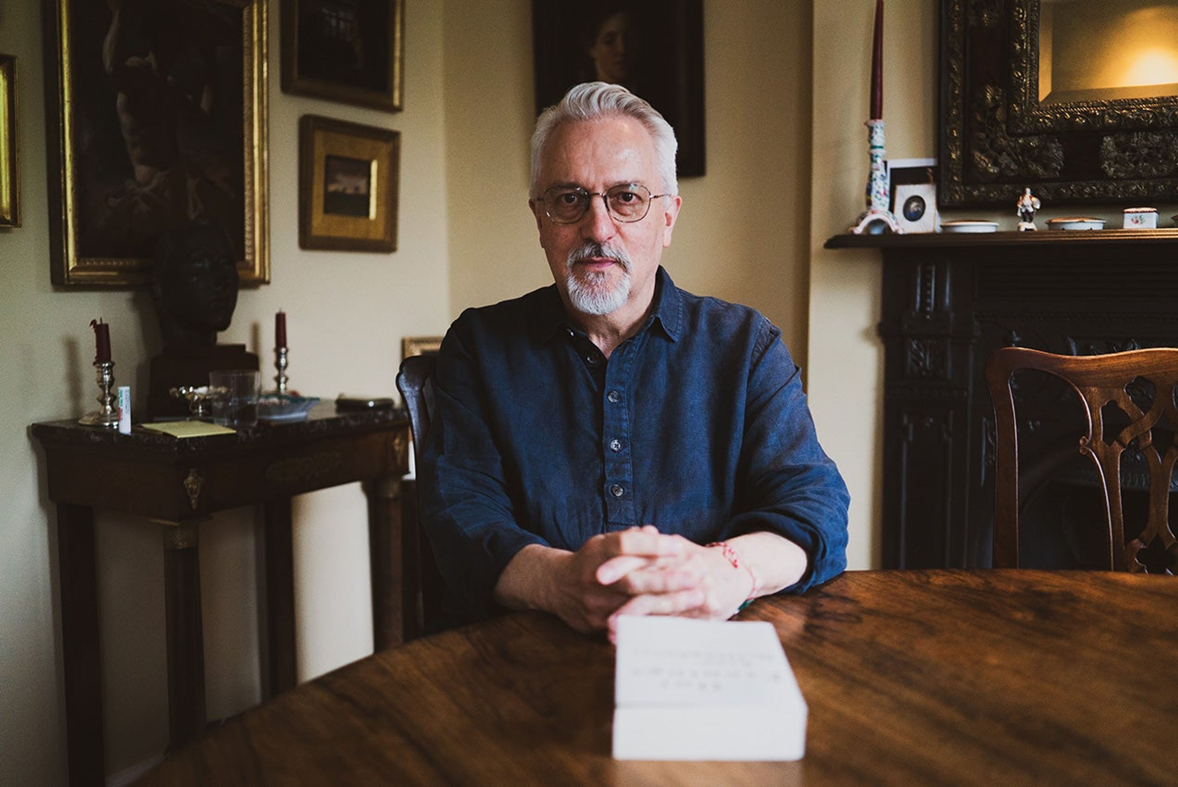 A photograph of Alan Hollinghurst sitting at table in a blue shirt