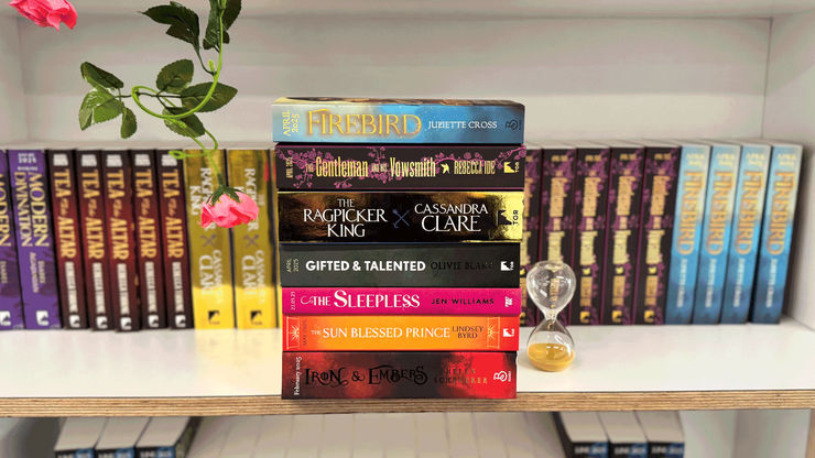 A stack of fantasy books sit on a bookshelf, in front of many other books blurred in the background. A rose dangles in front of the books and a sand hourglass sits next to the books.