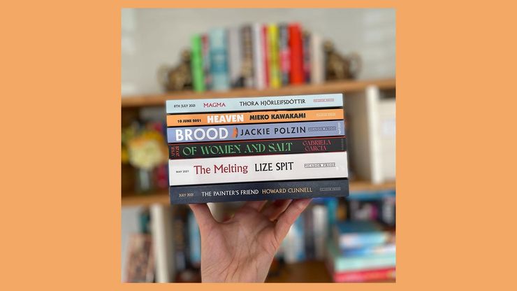 A photograph of a hand wielding a stack of books in front of a blurred out bookcase, on an orange background.