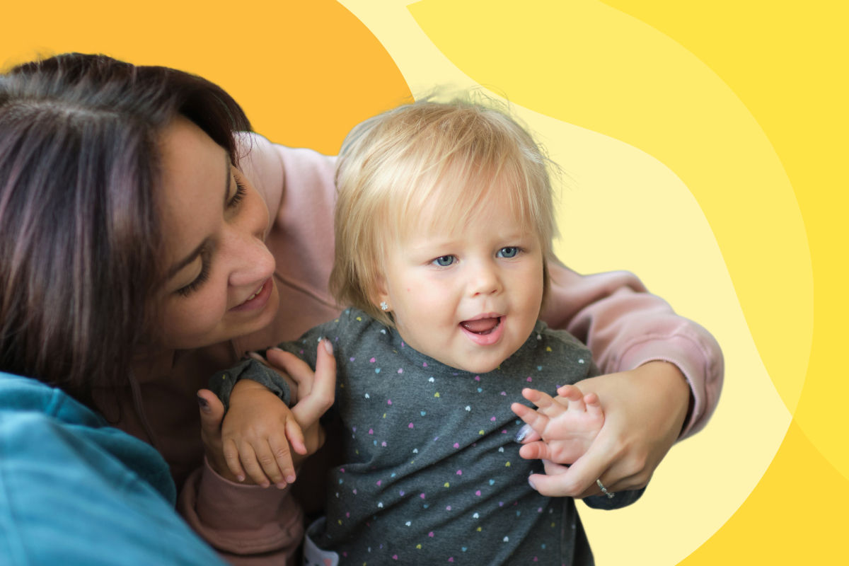 A woman holds her child up to stand from behind while he grabs her hands and smiles.