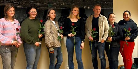 7 personer med hver sin rose står foran en vegg. Foto