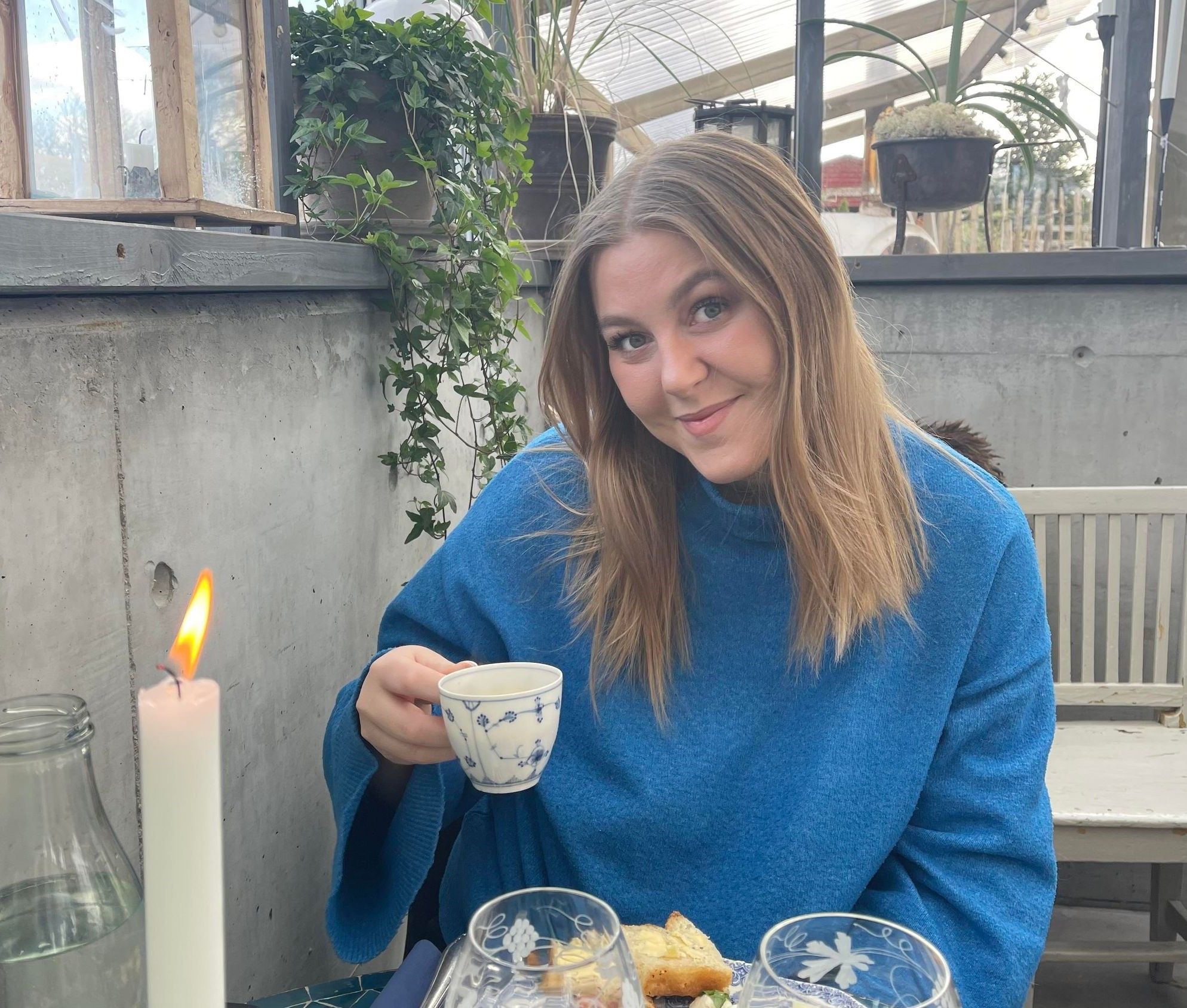 Bilde av Caroline Madsen. Kvinne med blondt hår, som smiler mot kamera. Hun har på seg en blå genser og holder en kopp. Hun sitter ved noe som ser ut som et cafébord, med mat og lys på bordet. 