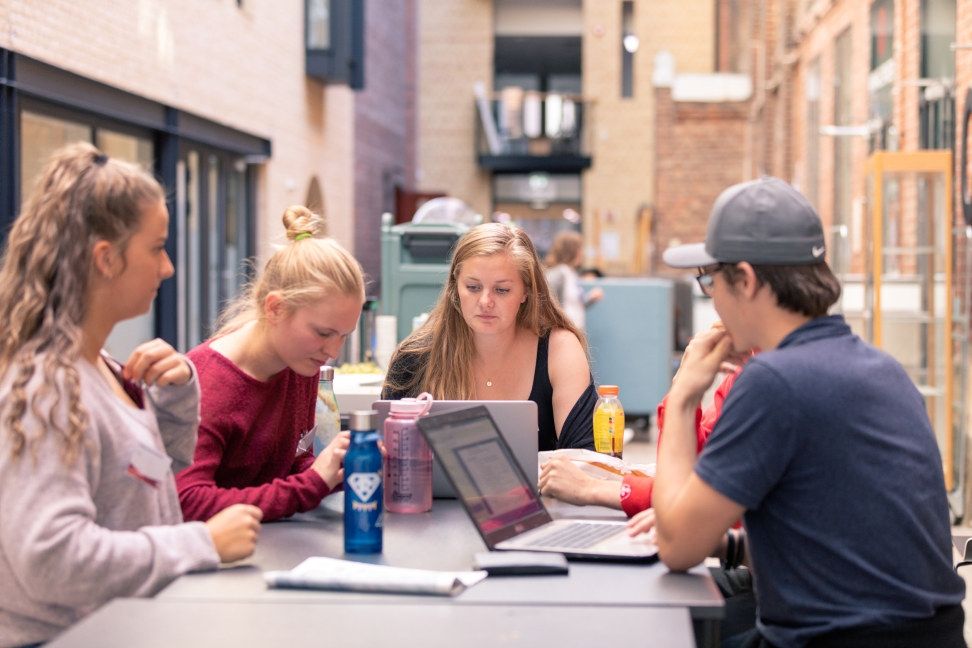 Fire studenter som sitter rundt et bord, med datamaskiner og bøker åpne. Alle ser vekk fra kamera. 