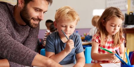 Smilende lærer so sitter ved pulten til to elever i småskolen og viser dem noe i bøkene. Illustrasjonsfoto