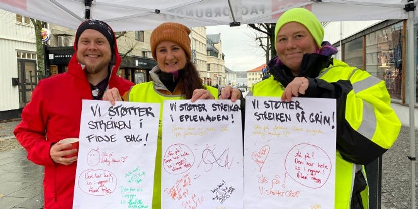 Vi ser bilde av tre streikende på stand med streikevester og plakater.