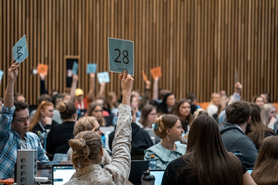 Flere mennesker i en møtesal. Deltakerne stemmer ved blå og oransje stemmesedler. Oversiktsbilde. Foto.