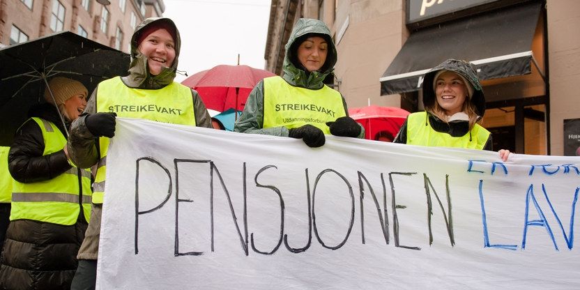 Smilende kvinner i regntøy og streikevester holder et banner der man kun kan lese følgende del av parolen " Pensjonen lav" 