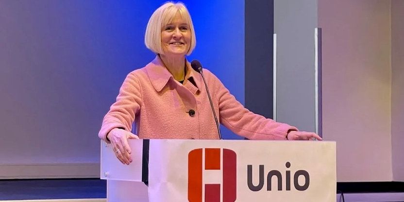 Ragnhild Lied står foran en talerstol på Unios konferanse. Hun har kort hvitt hår med pannelugg og har på seg en rosa dressjakke. Hun holder talestoler med begge hender og ser utover forsamlingen og smiler.
