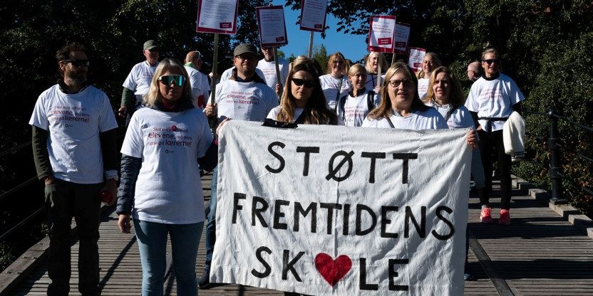 En gruppe med 15-20 lærere i streikeskjorter viser frem et banner med teksten "STØTT FREMTIDENS SKOLE" under fjorårets lærerstreik. O-en i teksten er formet som et hjerte.
