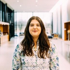Bilde av Karen Joanna Melby Myhre. Kvinne med langt, brunt hår smiler mot kamera. Hun har på en lyseblå kjole, med blonder og blomster. Foto. Fotograf: Adrian Castillo.