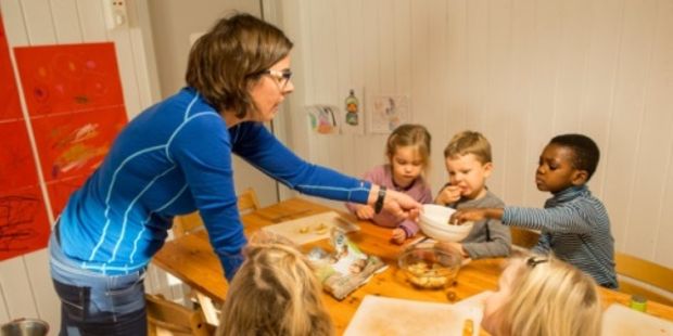 kvinnelig barnehagepersonal står med en skål i hånden som et barn tar fra, mens vi ser fire andre barn som sitter ved bordet