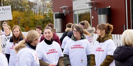 Flere kvinner står i streike-t-skjorter med påskriften Streik i barnehagen. I bakgrunnen ser ved en bygning der det står  Store Tune Gård barnehage