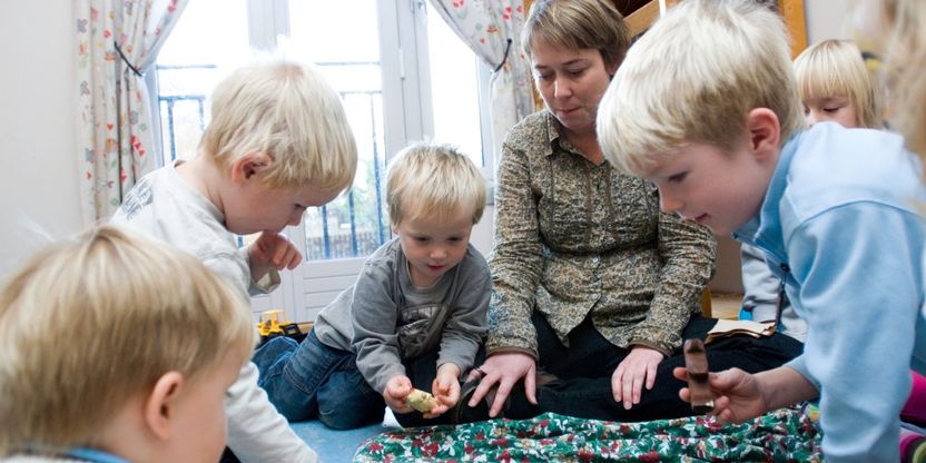 Barn sitter rundt en kvinnelig barnehagelærer på gulvet i en barnehage og leker.