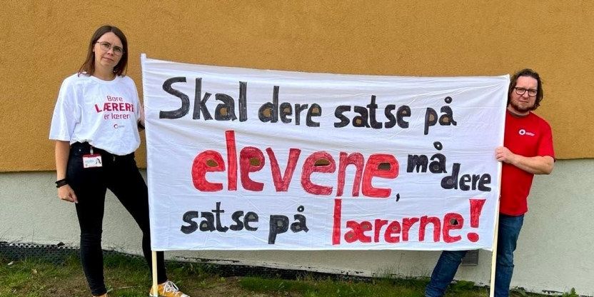 Bilde av Gunhild Grindjordet og Roar Busterud: De står mot en vegg med en banner mellom seg som sier: Skal dere satse på elevene, må dere satse på lærerne!