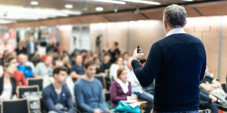 En mann står med ryggen mot fotograf og underviser en mengde mennesker i et auditorium.