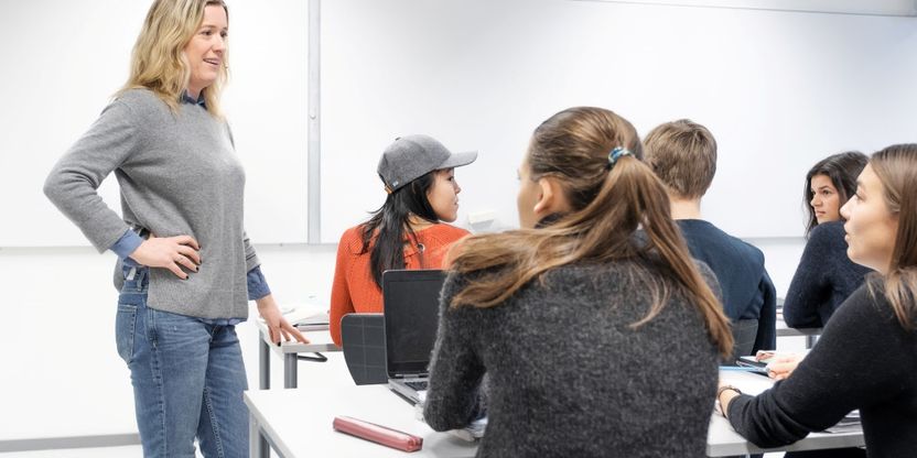 Kvinnelig lærer står ved pulten til  flere ungdommer og snakker med dem. De er i et klasserom.