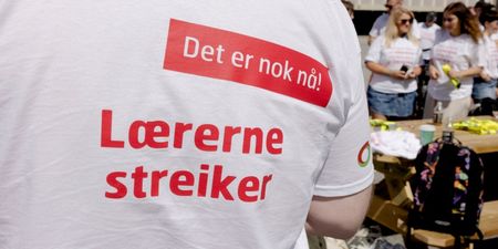 Person står med ryggen mot kamera med streike t-skjorte på seg. I bakgrunnen ses andre streikende. 