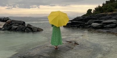 Dame som står på en stein i fjæra med grønn kjole som holder en gul paraply og ser ut over havet