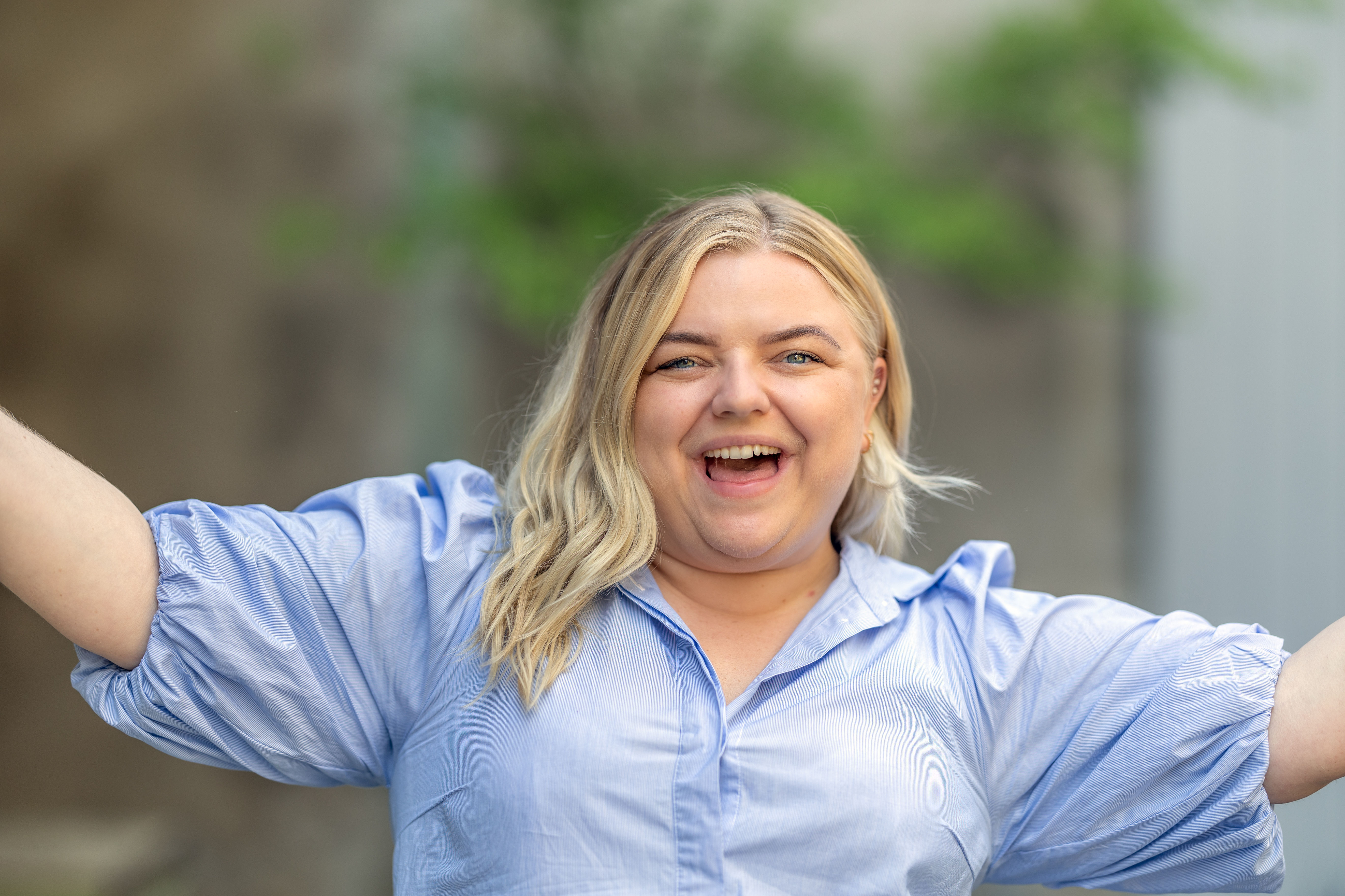 Blond kvinne smiler stort med hendene ut til siden. Hun har på en blå skjorte og bakgrunnen er ute av fokus. Foto. 