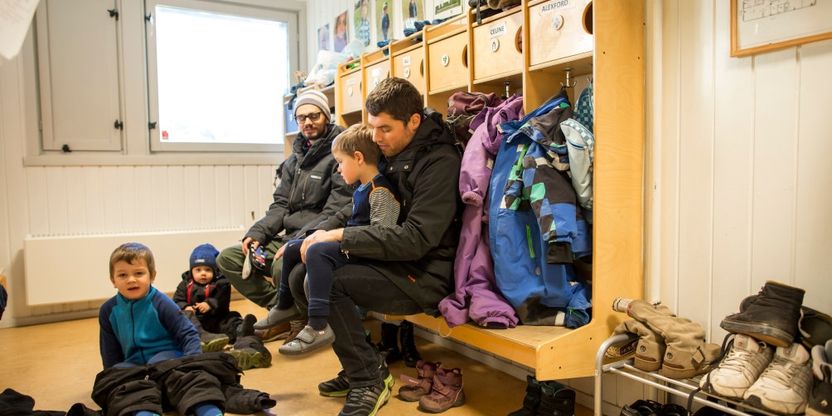 Et barnehagebarn  sitter på fanget til en voksen mann og blir kledd på yttertøy i garderoben i en barnehage. I bakgrunnen  ser vi en annen voksen mann og  flere barn.