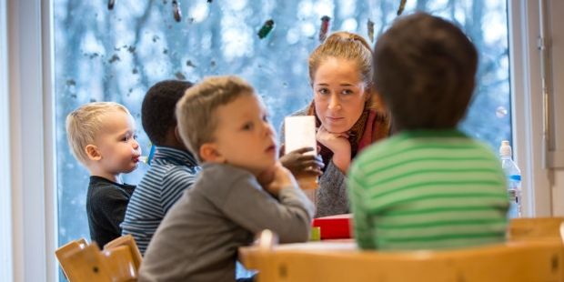 Bilde av barnehagelærer som sitter ved et bord sammen med flere barn.
