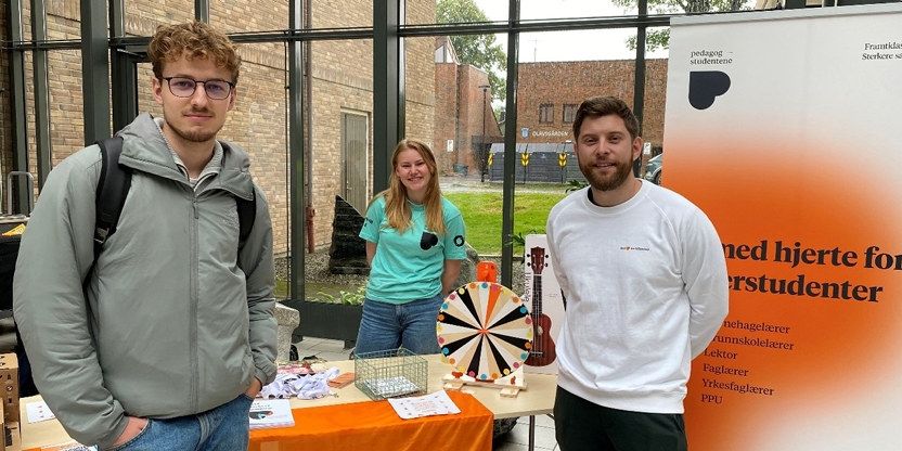 Tre studenter på stand, med en roll-up fra Pedagogstudentene i oransje og hvit til høyre i bildet.