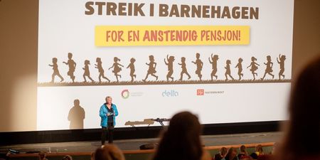 Person som står på en scene i et auditorium foran tilskuere. På skjerm i bakgrunn er streikelogo for barnehagestreiken 2022, vist.