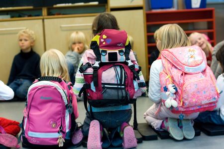 Barn som sitter i en ring på et gulv. Noen med ransel på. Foto. 