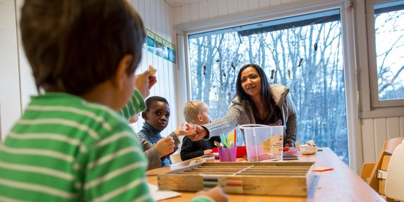 En kvinnelig barnehagelærer deler ut mat til tre barn som sitter ved et langbord inne i en barnehage. Det er vinterlig vær gjennom vinduet. 