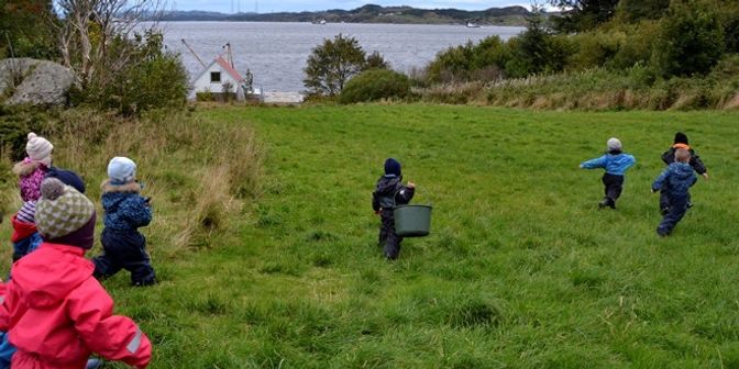 Barnehagebarn på en grønn slette eller svak hellende bakke, ned mot havet. 