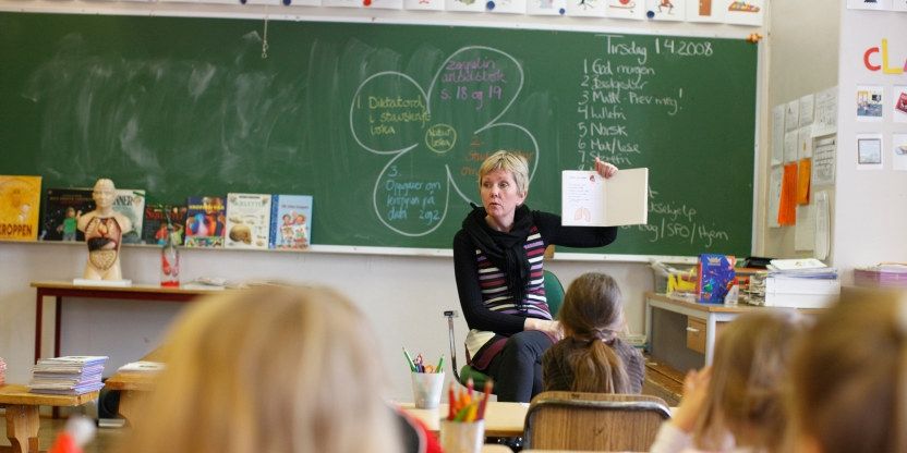 En kvinnelig lærer i klasserommet holder opp et ark foran en klasse med yngre barn. 