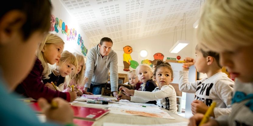 En mannlig barnehagelærer som lener seg over et langbord med åtte-ni barn i 3-4 årsalderen. 