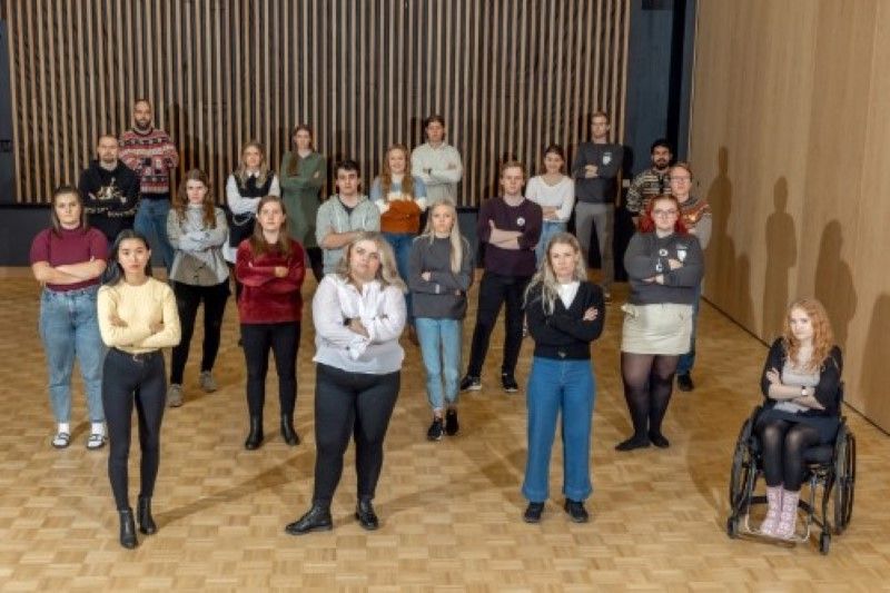 Gruppebilde av 21 mennesker som står på koronavennlig avstand til hverandre i en sal med tregulv og en vegg i tre den andre med trespiler på sort bakgrunn. Alle har armene i kors og ser alvorlig ut. 