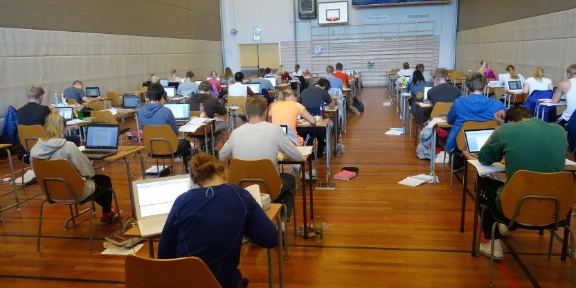 En gymsal som viser videregående elever som tar eksamen. De sitter på rekke og rad med ryggen vendt mot kamerat. På pultene står det PCer og oppslåtte bøker. 