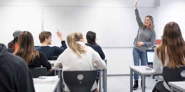 Bilde av elever som sitter med ryggen til ved pultene sine i et klasserom. En lærer står foran elevene og snakker til klassen.