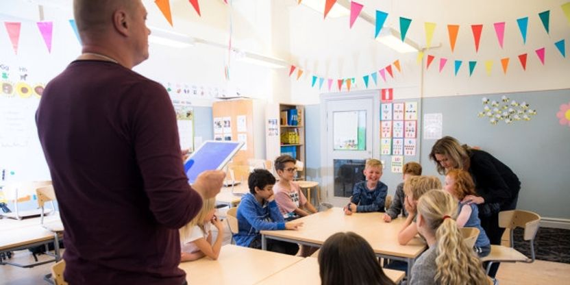 Foto av mannlig lærer som står foran barneskoleklasse ved pulter. Girlandere i taket. 