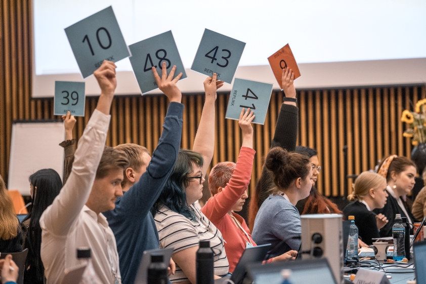 Bilde av delegater på landsmøte, som strekker stemmeskilt i været. 