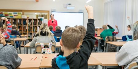 Bilde av elever sett bakfra hvor et par som rekker opp hånda. I forgrunnen står en l'ærer. Denne personen er litt utydelig og fokus er på elevene.