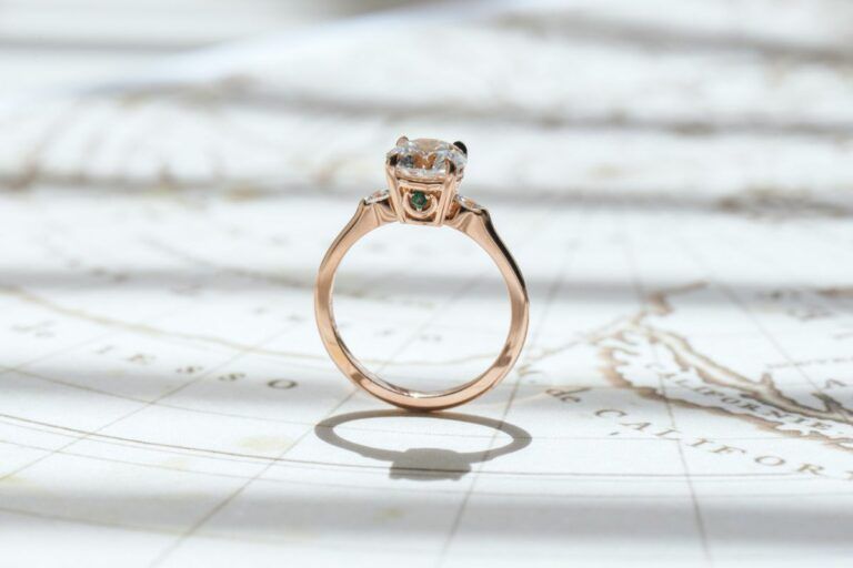 A round-cut rose gold engagement ring featuring a compass logo under the center stone. A tiny rhodolite garnet and emerald are on each side, placed atop a crescent.