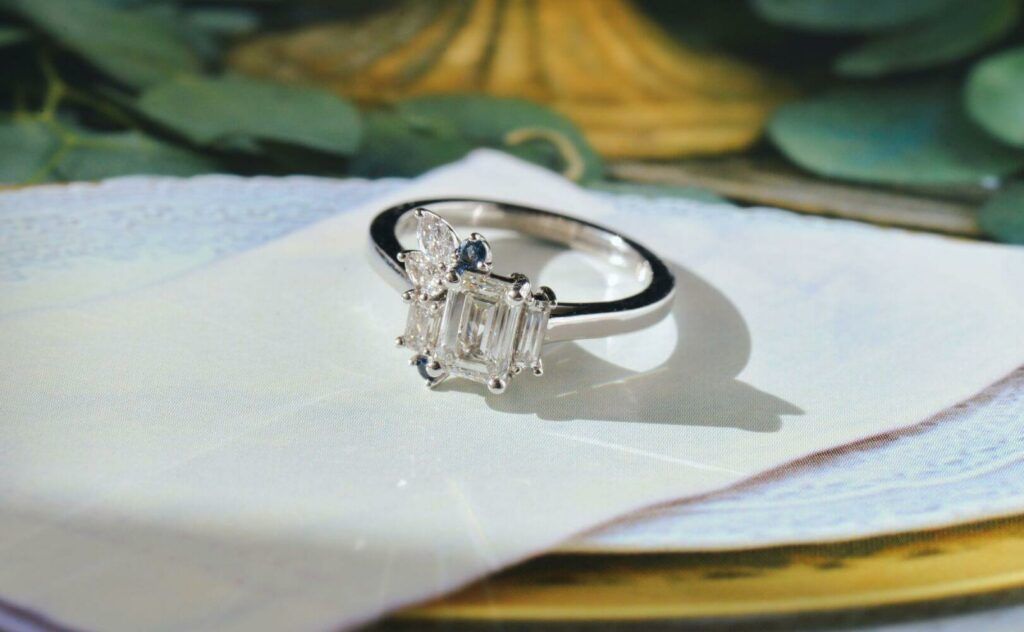 A lab-grown emerald-cut diamond ring with two straight baguette cuts (one longer than the other). Atop the shorter baguette sits a pear and marquis diamond, and a round blue sapphire.