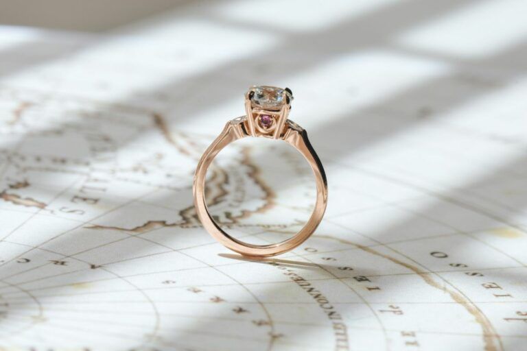 A round-cut rose gold engagement ring featuring a compass logo under the center stone. A tiny rhodolite garnet and emerald are on each side, placed atop a crescent.