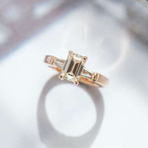 A 14k rose gold engagement ring featuring an emerald cut center stone and tapered baguette side stones.