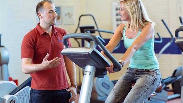 vrouw op fitnessfiets met begeleider naast zich bij de fysio