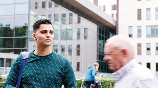 jongere man en ouderen man voor gebouw met bellende dame op de achtergrond bij haar fiets