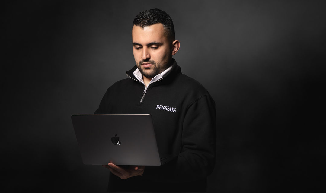 A person wearing a black Perseus Creative Studio branded quarter-zip top is adjusting a large lens on a camera body in an indoor setting, highlighting the process of setting up professional filming equipment.