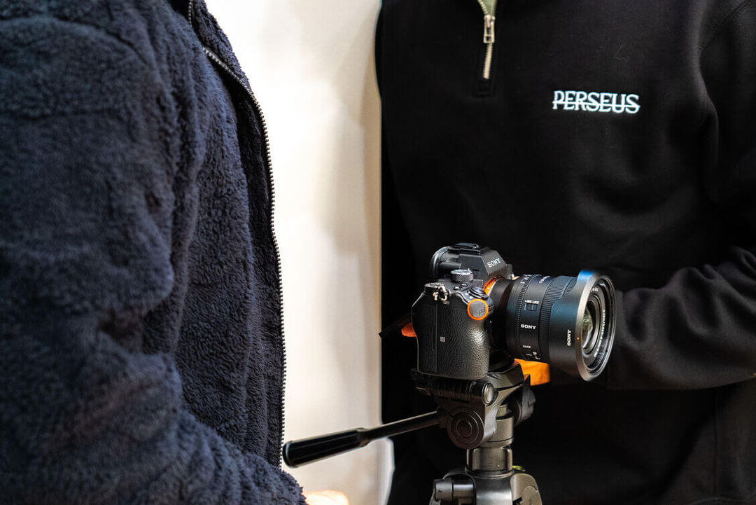 Two people filming indoors with a Sony mirrorless camera on a tripod; the cameraman is wearing a black Perseus Creative Studio branded quarter-zip sweater, demonstrating a professional video production setup.