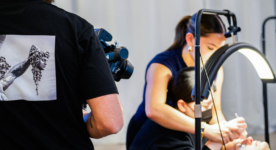 Behind-the-scenes shot of a videographer wearing a black t-shirt with a Medusa/Perseus graphic print on the back, filming a beauty treatment or service under a bright ring light.