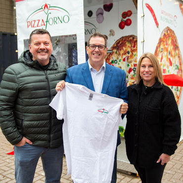 Three people standing in front of a PizzaForno machine holding a PizzaForno t-shirt