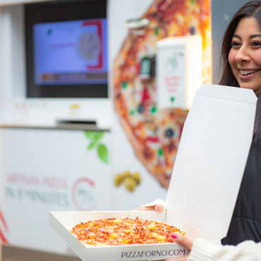 A girl is holding a PizzaForno box with a Hawaiian pizza inside it, she stands in front of a pizza vending machine