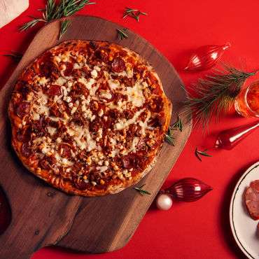 A Meat Lovers pizza on a brown cutting board with meat and vegetables beside it. 